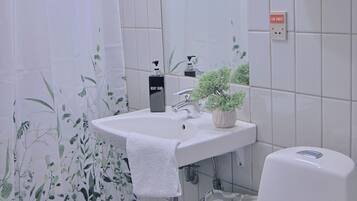 Un baño con lavamanos blanco, inodoro, cortina de ducha, estante con artículos de aseo y una planta en maceta.