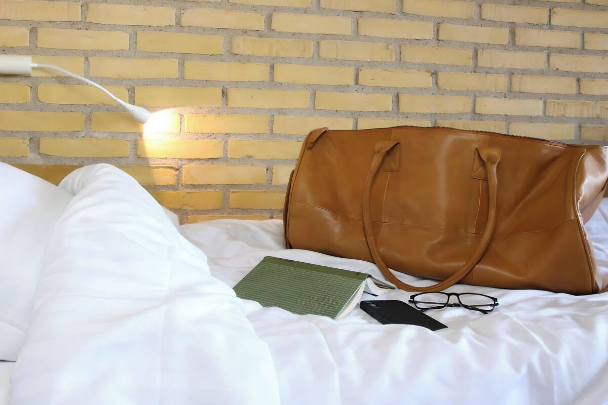 A bed with white bedding, a brown leather bag, a green notebook, and a pair of glasses on it.