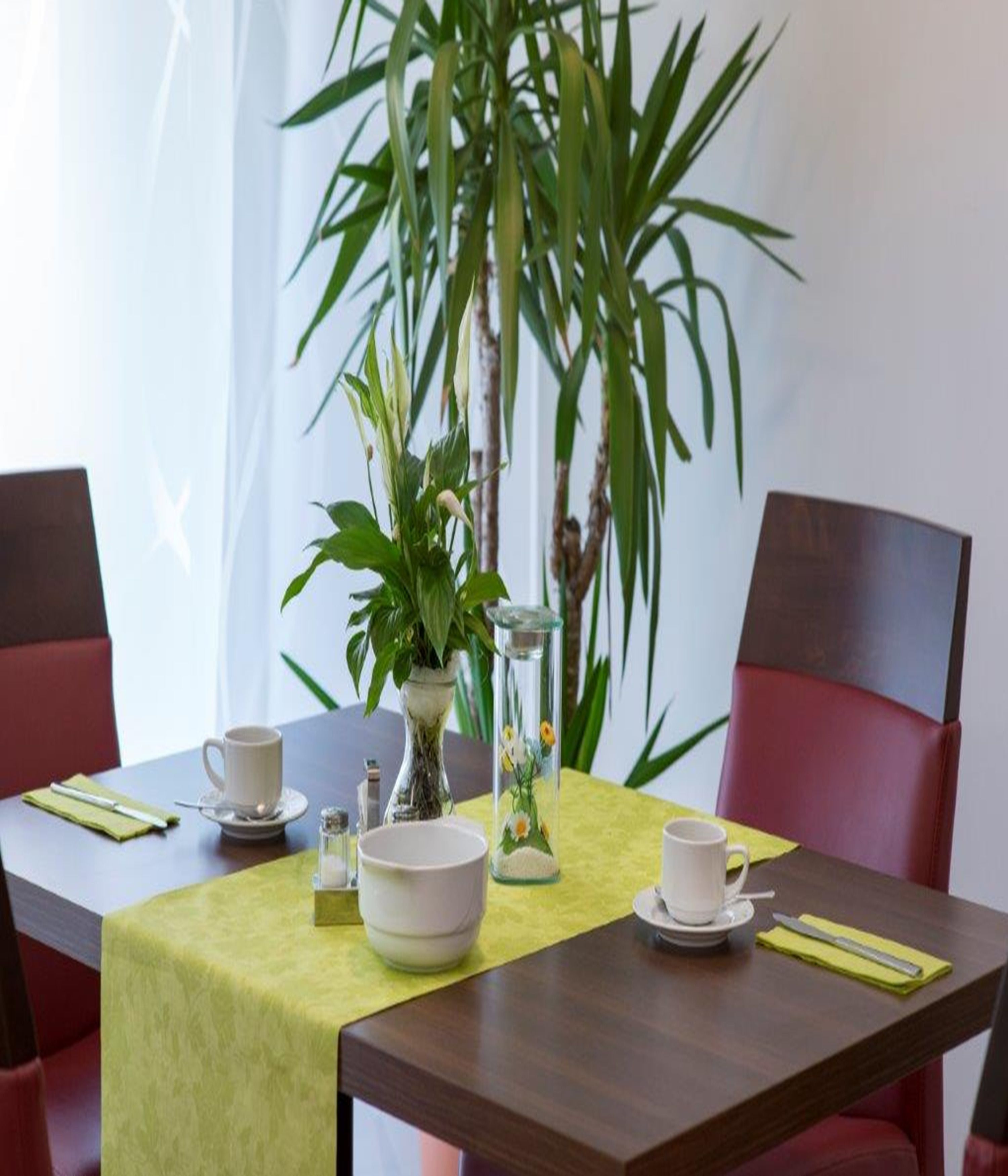 A dining table with a green tablecloth, white bowls, a white cup, and a glass vase with flowers. There are red chairs around the table.