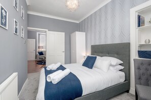A bedroom with a large bed, a grey upholstered headboard, a white wardrobe, and a grey armchair. - No 10 - by StayDunfermline (Dunfermline)