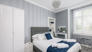 Un dormitorio moderno con un cabecero tapizado en gris, un armario blanco, una mesita de noche con lámpara y una ventana con persianas.