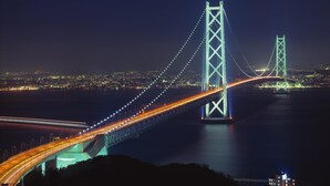 Vista nocturna de un gran puente colgante con cables y torres iluminados, que se extiende sobre una masa de agua con luces de ciudad al fondo.