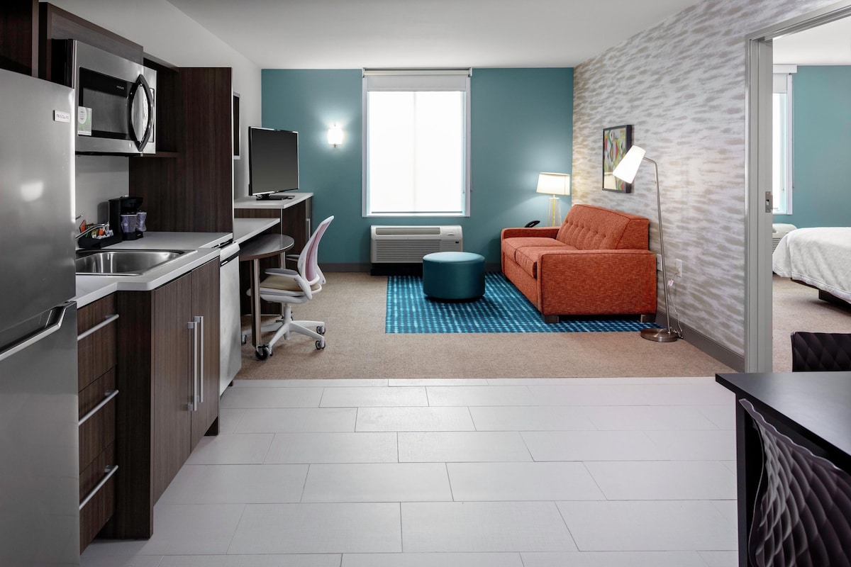 A compact hotel room with a kitchenette, a single bed, a sofa, a desk, and a chair.