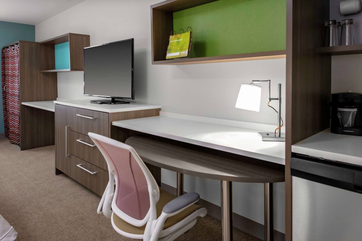 A modern hotel room with a desk, chair, television, and a coffee machine.