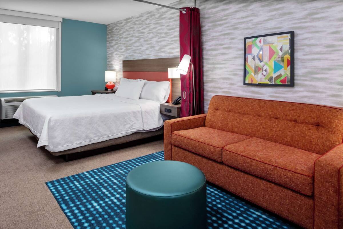A hotel room with a bed, a sofa, a nightstand, a lamp, a wall art piece, and a patterned rug.