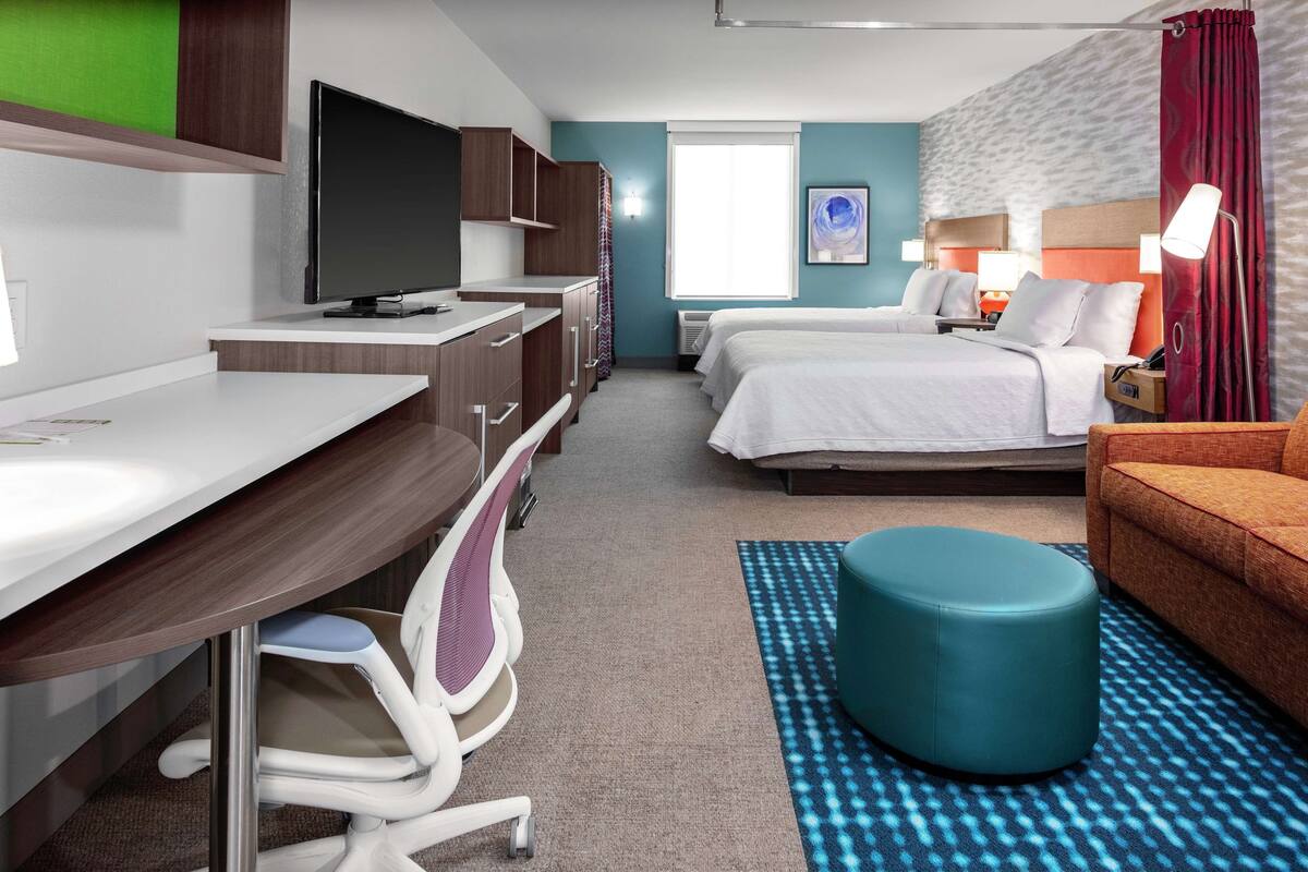 A hotel room with a desk, two beds, a TV, a sofa, and a patterned rug.