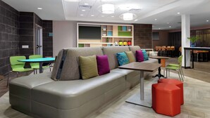 A modern lounge area with a large sofa, a small table, and a red ottoman. There are chairs and a bar area in the background. - Home2 Suites by Hilton North Charleston-University Blvd (North Charleston)
