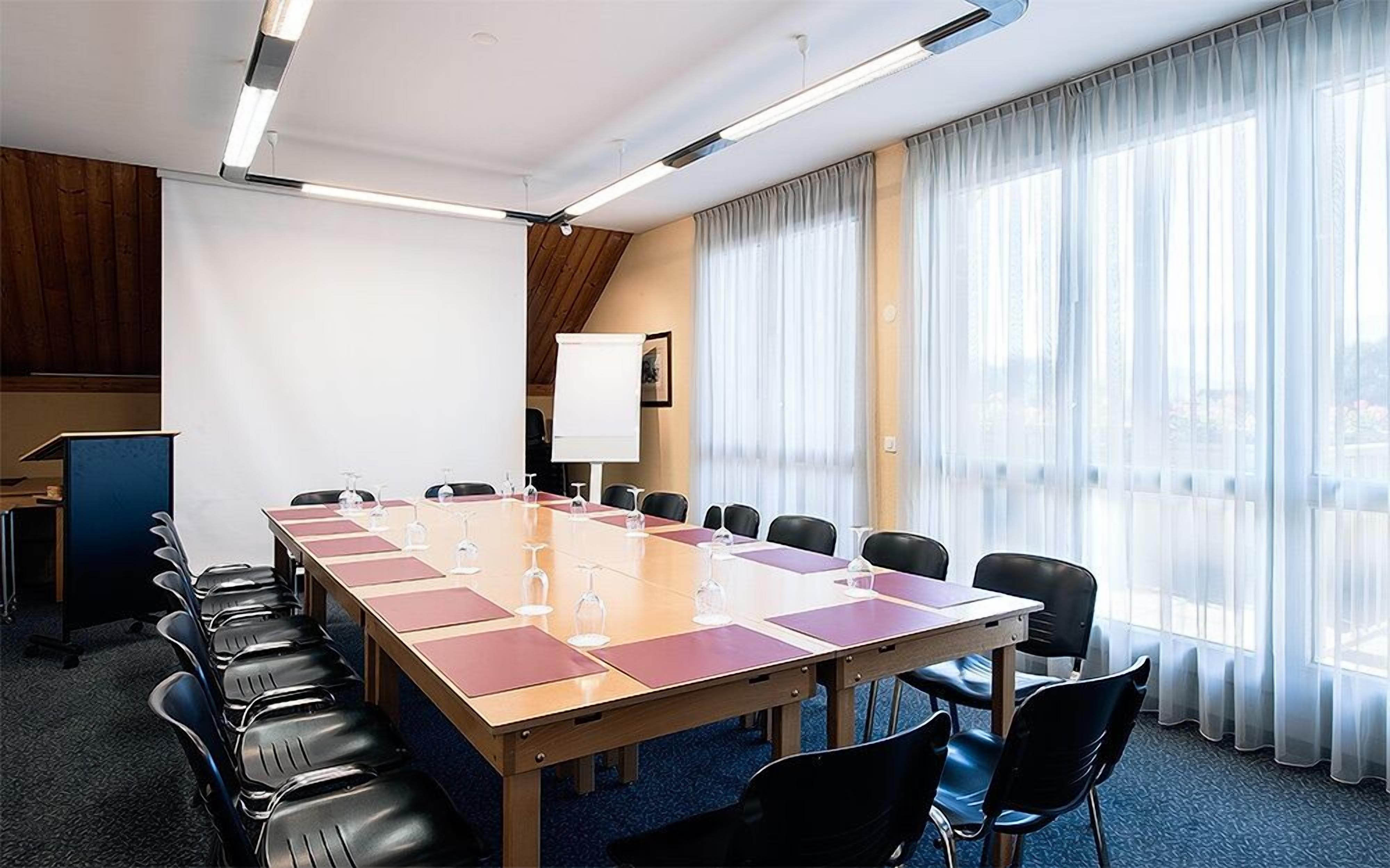 Ein Konferenzraum mit einem langen Tisch, Stühlen, einer Whiteboard-Tafel und großen Fenstern mit leichten Vorhängen.