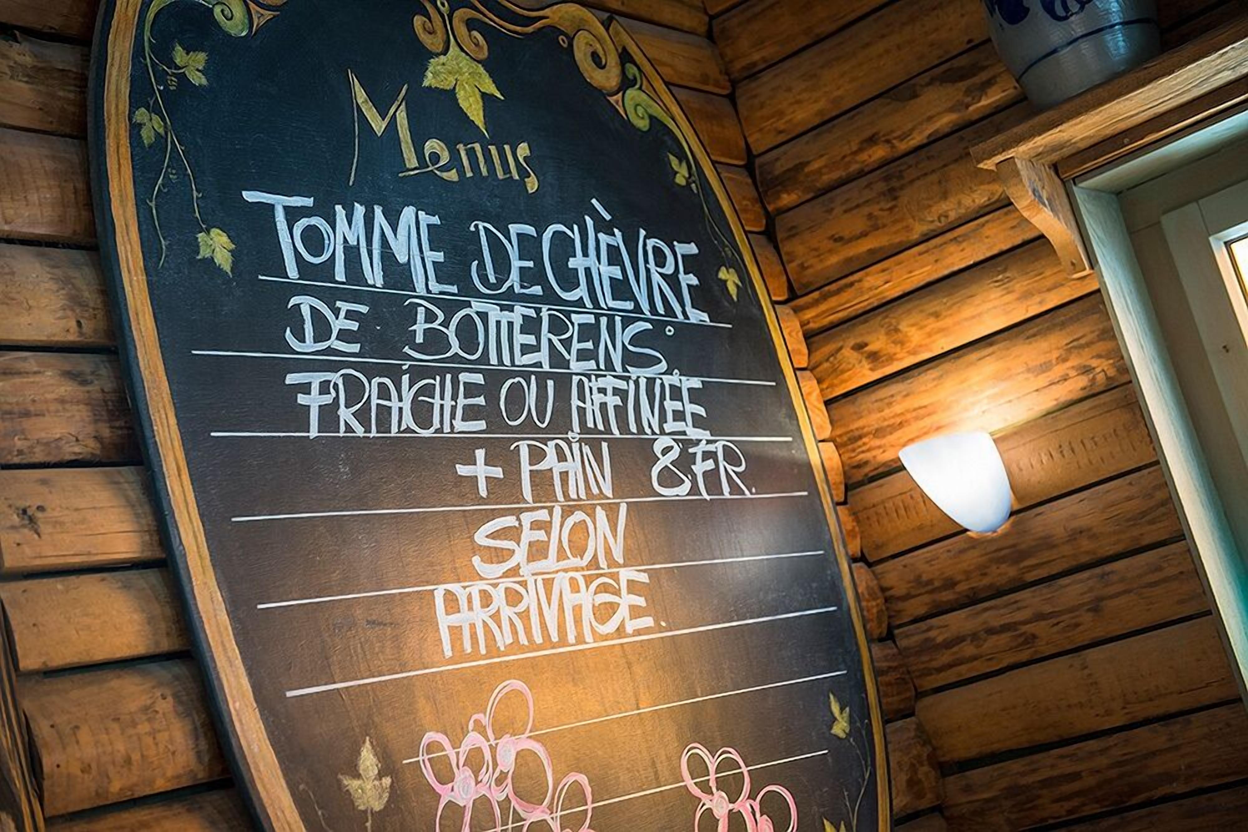 Un menú en pizarra dentro de un espacio de madera, que enumera productos como 'Tomme de chèvre de Botterens' y 'pain & fr'.