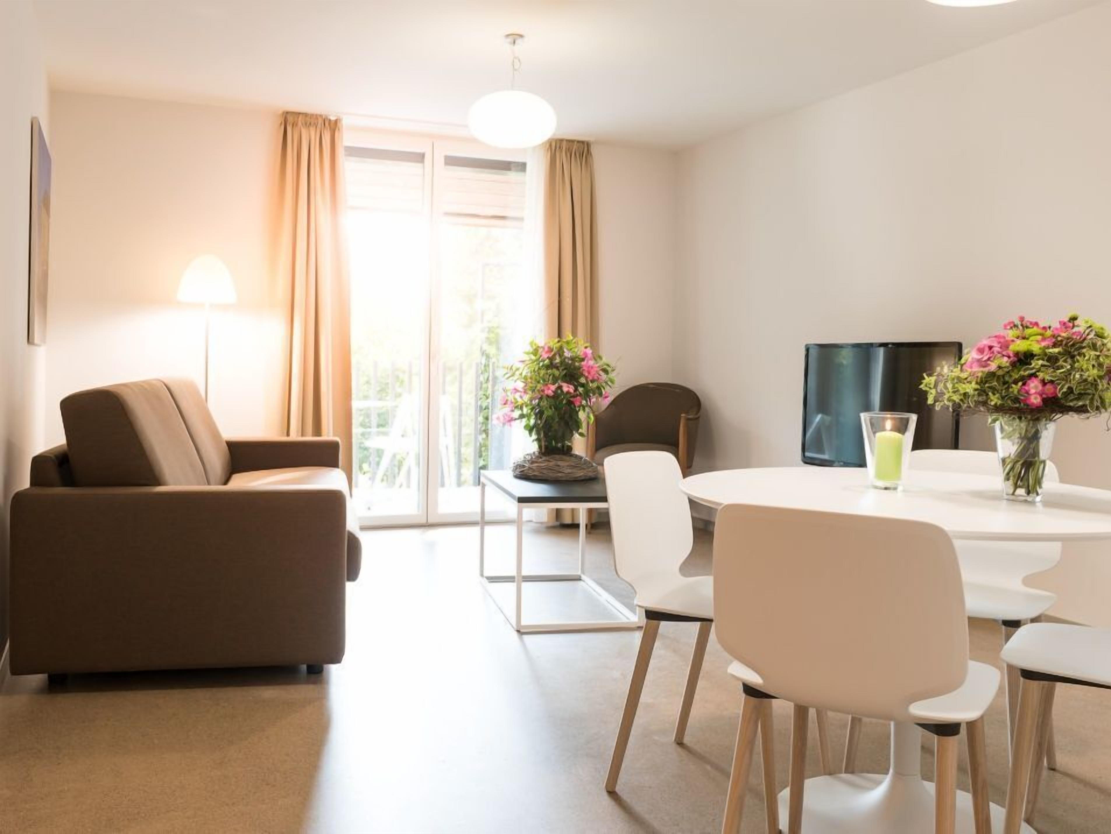 A modern living room with a sofa, chairs, a dining table, a TV, and a balcony with curtains.