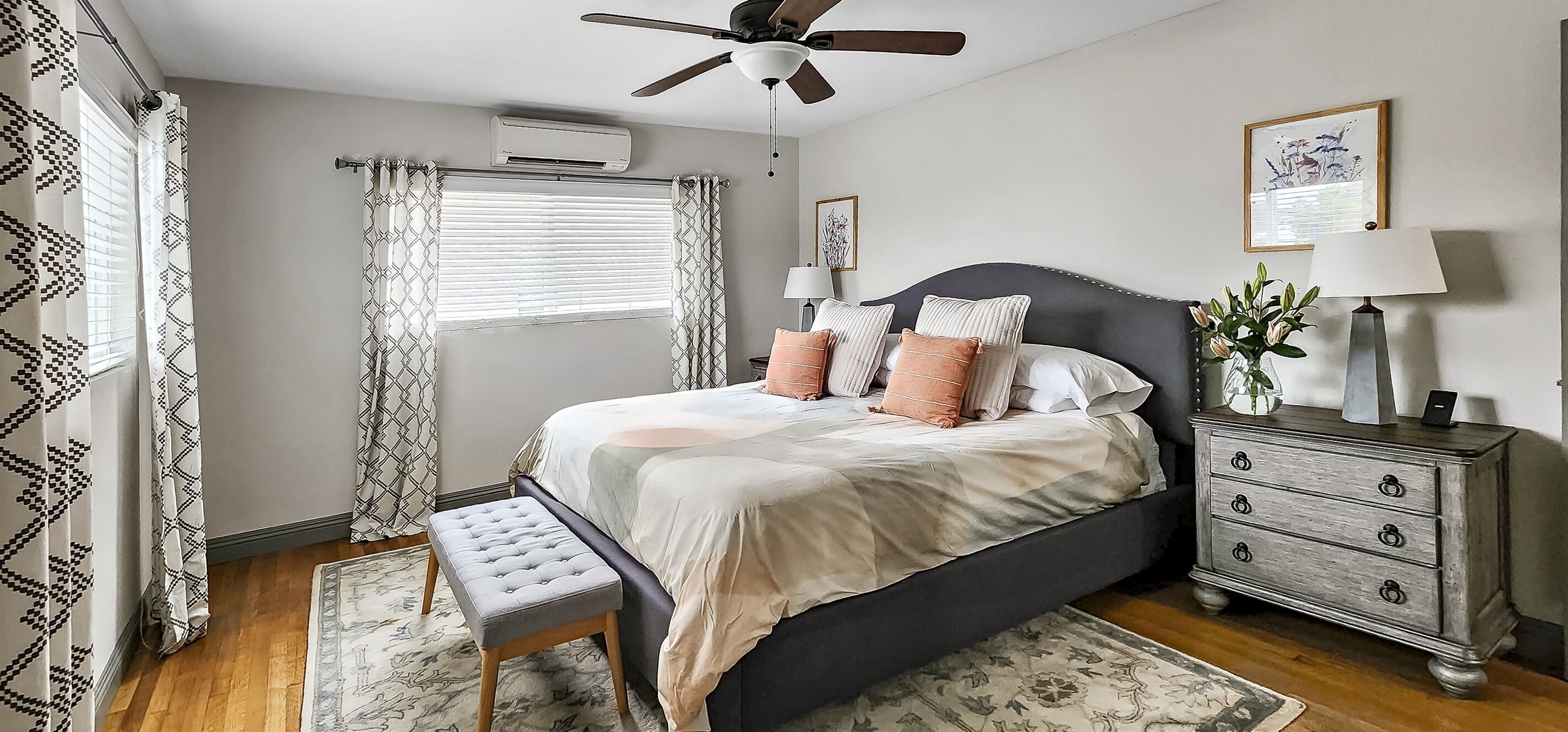 Une chambre à coucher comprenant un lit, des tables de chevet, un banc, un ventilateur de plafond, un climatiseur et une fenêtre avec des rideaux.