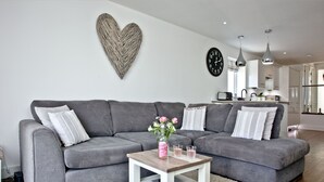 Ein modernes Wohnzimmer mit einem grauen Ecksofa, einem kleinen Holztisch mit einer Vase und Gläsern, einem weißen Teppich und einer Küche im Hintergrund.