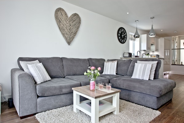 A modern living room with a grey sectional sofa, a small wooden coffee table with a vase and glasses, a white rug, and a kitchen in the background.