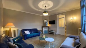 A living room with a blue sofa, a brown sofa, a small round table, a lamp, a fireplace, a TV, and a chandelier.
