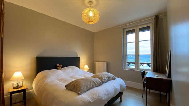 A bedroom with a large bed, bedside lamps, a desk, and a view of the sea through a window.