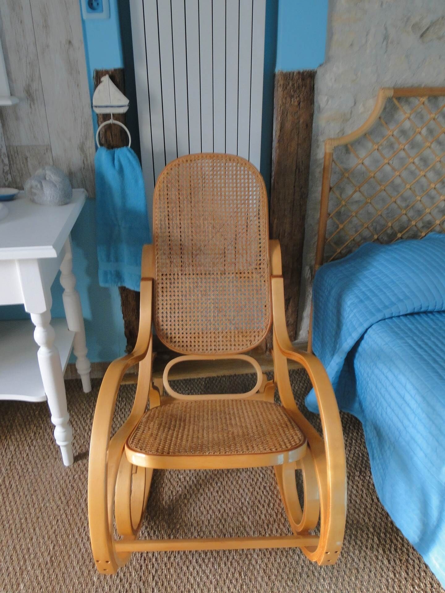 A wooden rocking chair with a woven seat and backrest, positioned in front of a bed with a blue quilt.