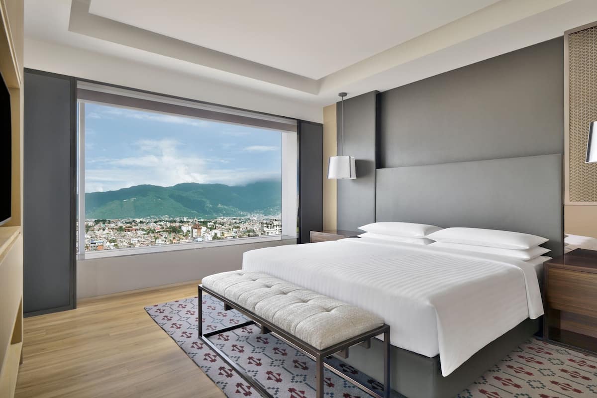 Ein modernes Hotelzimmer mit großem Fenster und Blick auf die Stadt, einem ordentlich bezogenen Bett, einer Bank und einem Nachttisch.
