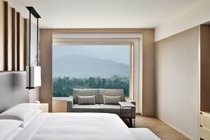 Una habitación de hotel moderna con una cama grande, un sofá, una mesita y vistas a un paisaje montañoso a través de un ventanal.