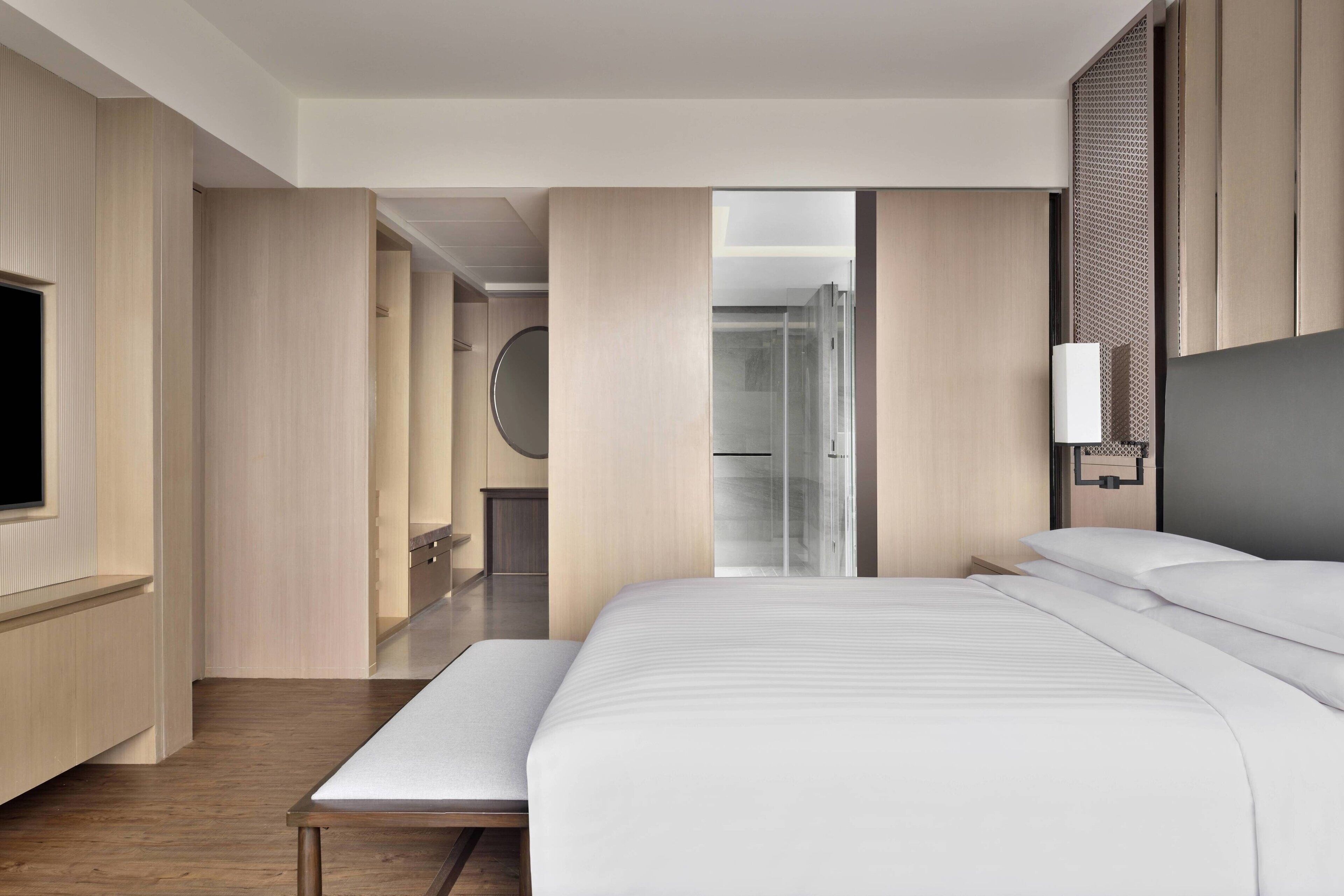 A modern hotel room with a large bed, a bedside table, a TV, and a bathroom visible through an open door.