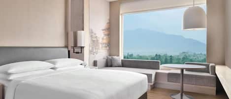 Habitación de hotel moderna con una cama grande, zona de estar con vistas a montañas y una mesa redonda.