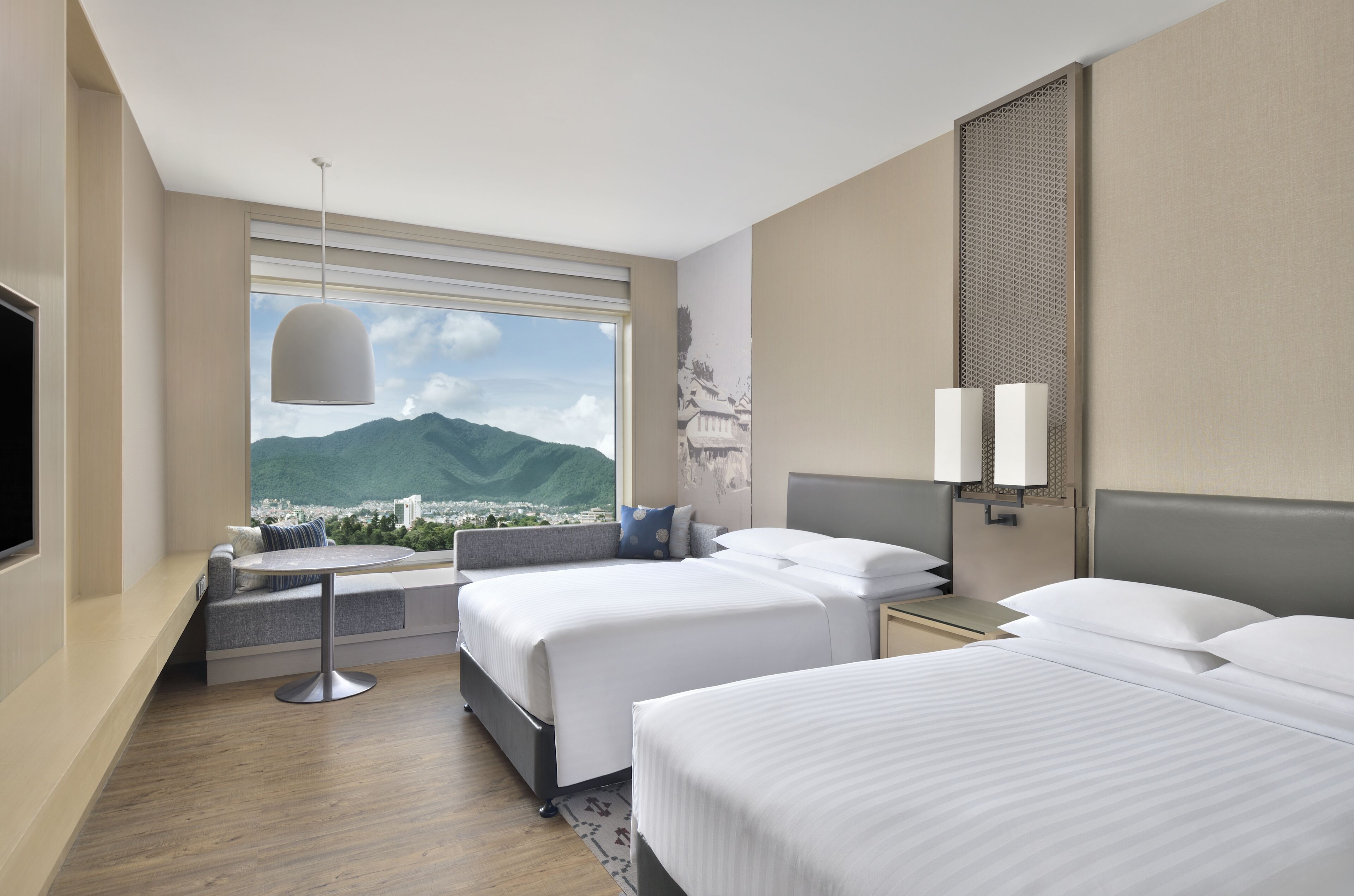 A hotel room with two beds, a seating area with a table, a large window with a mountain view, and a wall-mounted TV.