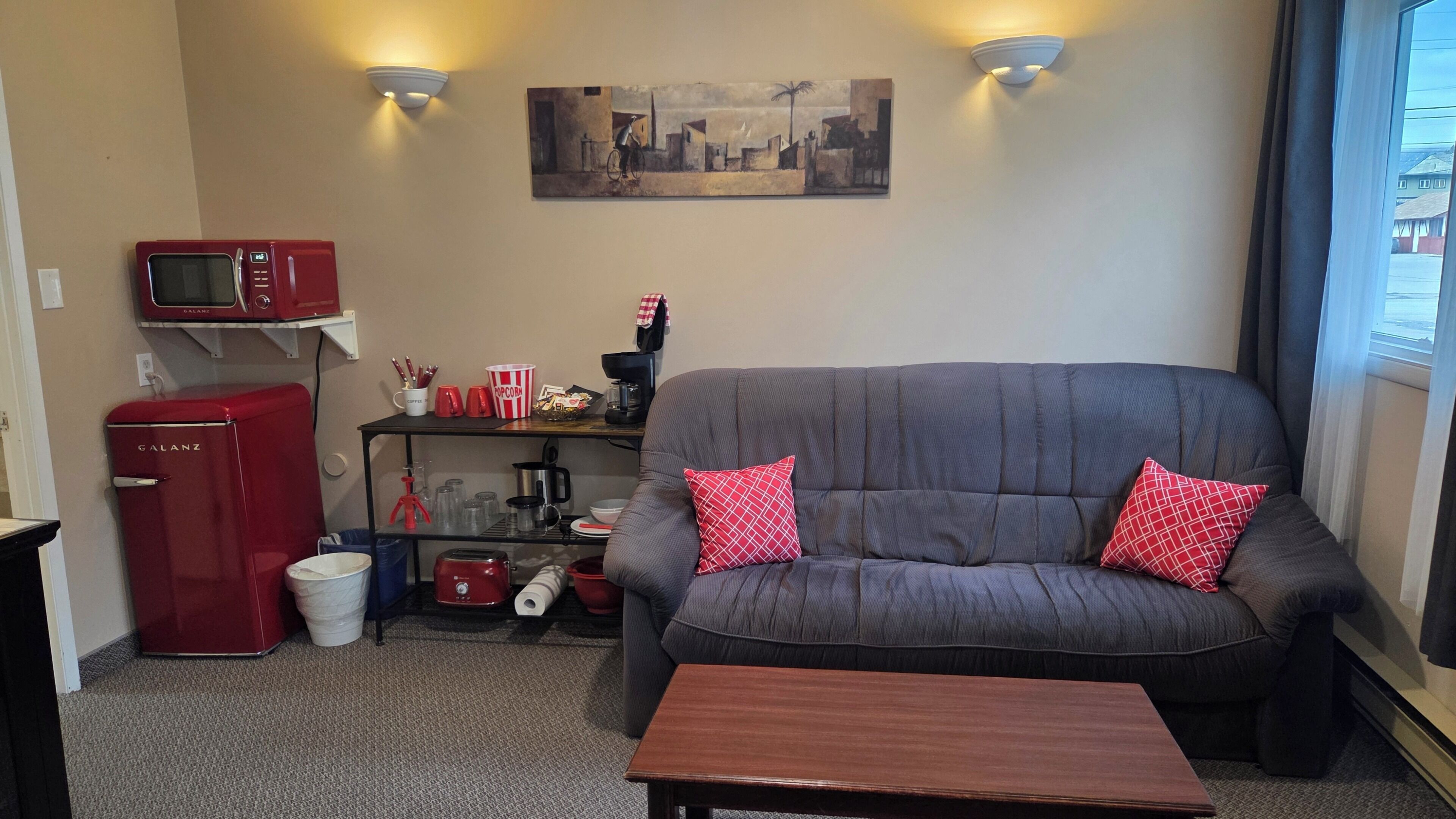 A living room with a gray sofa, two red pillows, a red microwave, a coffee maker, and a painting on the wall.