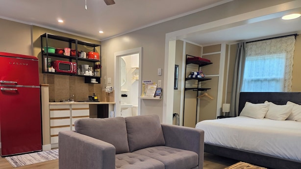 A compact hotel room with a grey sofa, a wooden coffee table, a bed with white linens, a red refrigerator, a kitchenette with shelves, and a window with curtains. - Lido Motel (Radium Hot Springs)