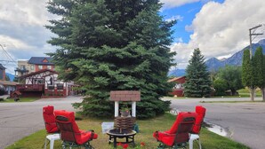 A cozy outdoor seating area with two red chairs and a small table, surrounded by a large evergreen tree and a brick planter with flowers. - Lido Motel (Radium Hot Springs)