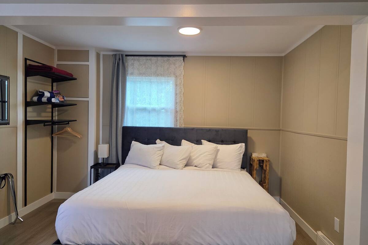 A neatly made bed with white linens and a dark headboard in a room with a wooden floor, a window with sheer curtains, and a shelf with folded clothes.