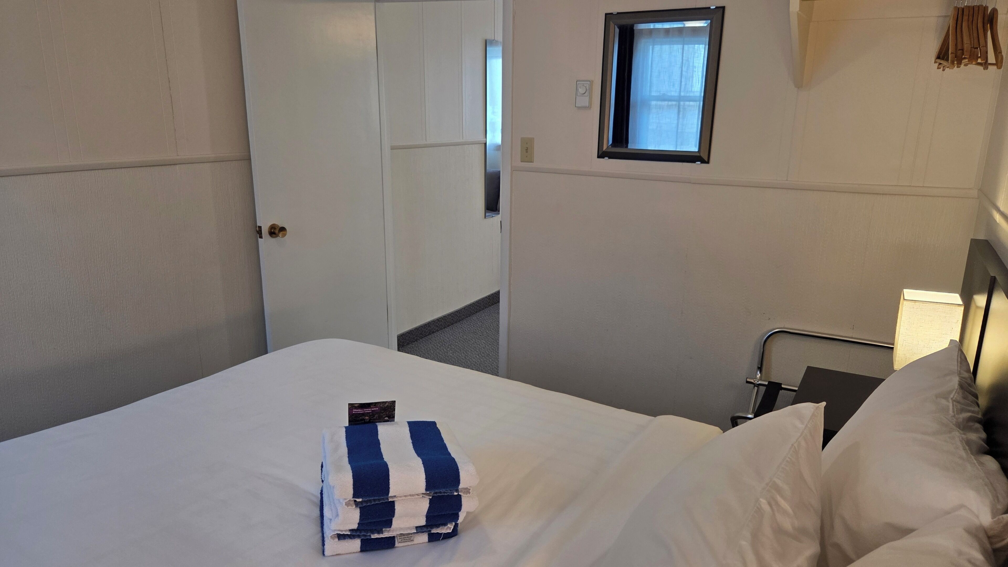 A hotel room with a bed, a window, a door, and a small window with curtains.