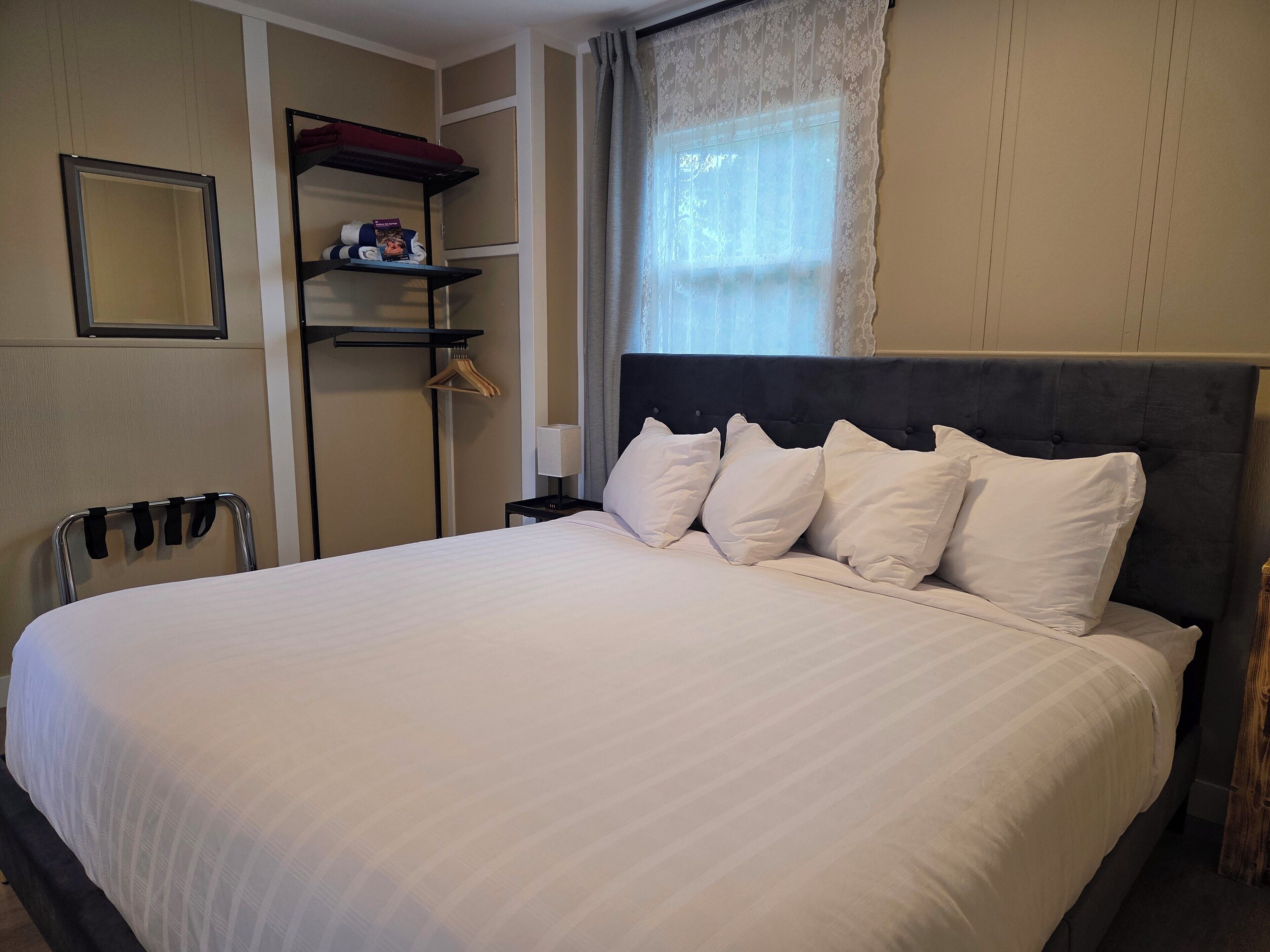 A neatly made bed with a dark headboard and white pillows, a bedside table with a lamp, and a wardrobe with hanging clothes.