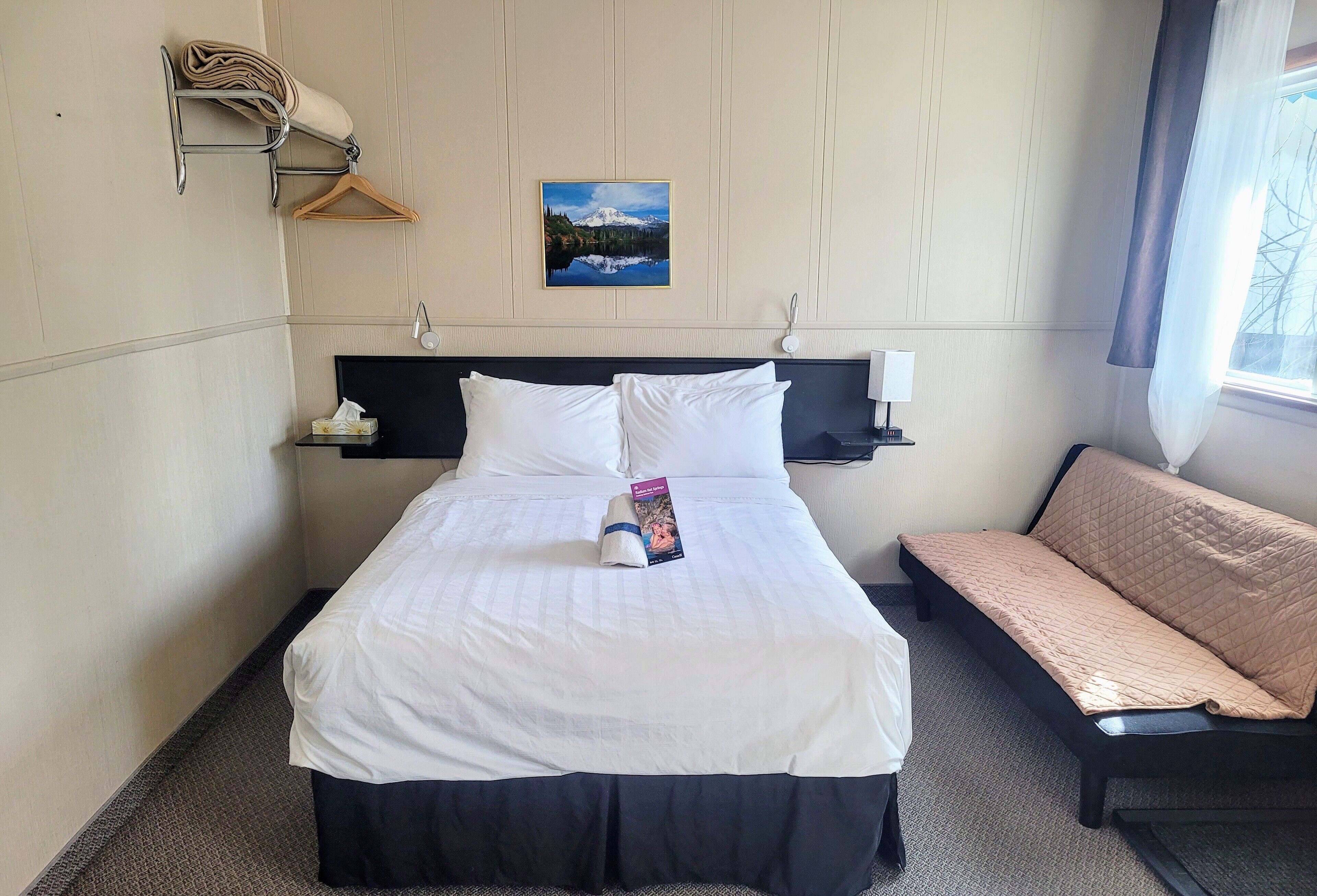 Une chambre d’hôtel comprenant un lit, une table de chevet, un porte-serviettes mural, un tableau accroché au mur et une fenêtre avec des rideaux.