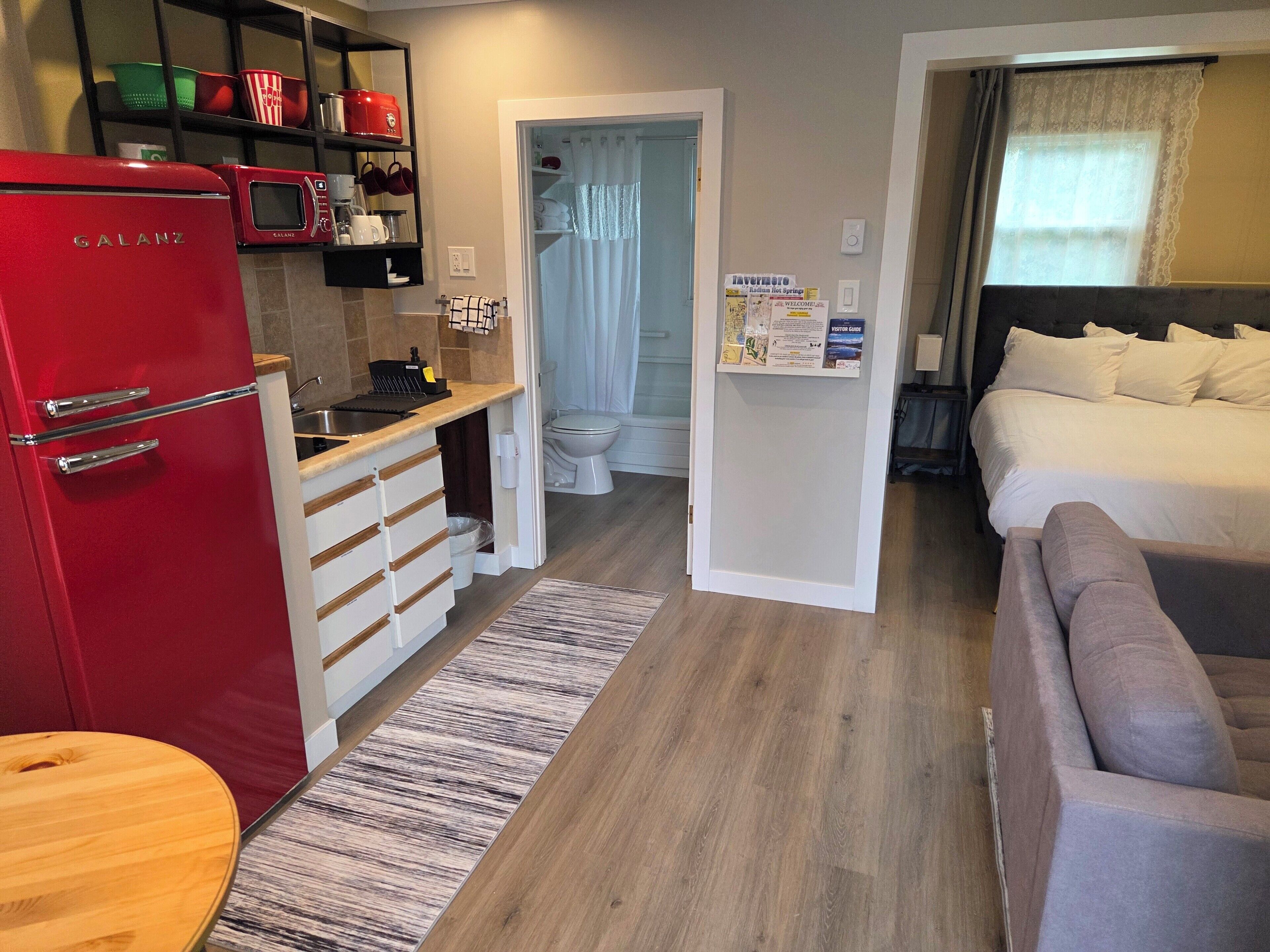 A compact hotel room with a kitchenette, a bed, a bathroom, and a small seating area.