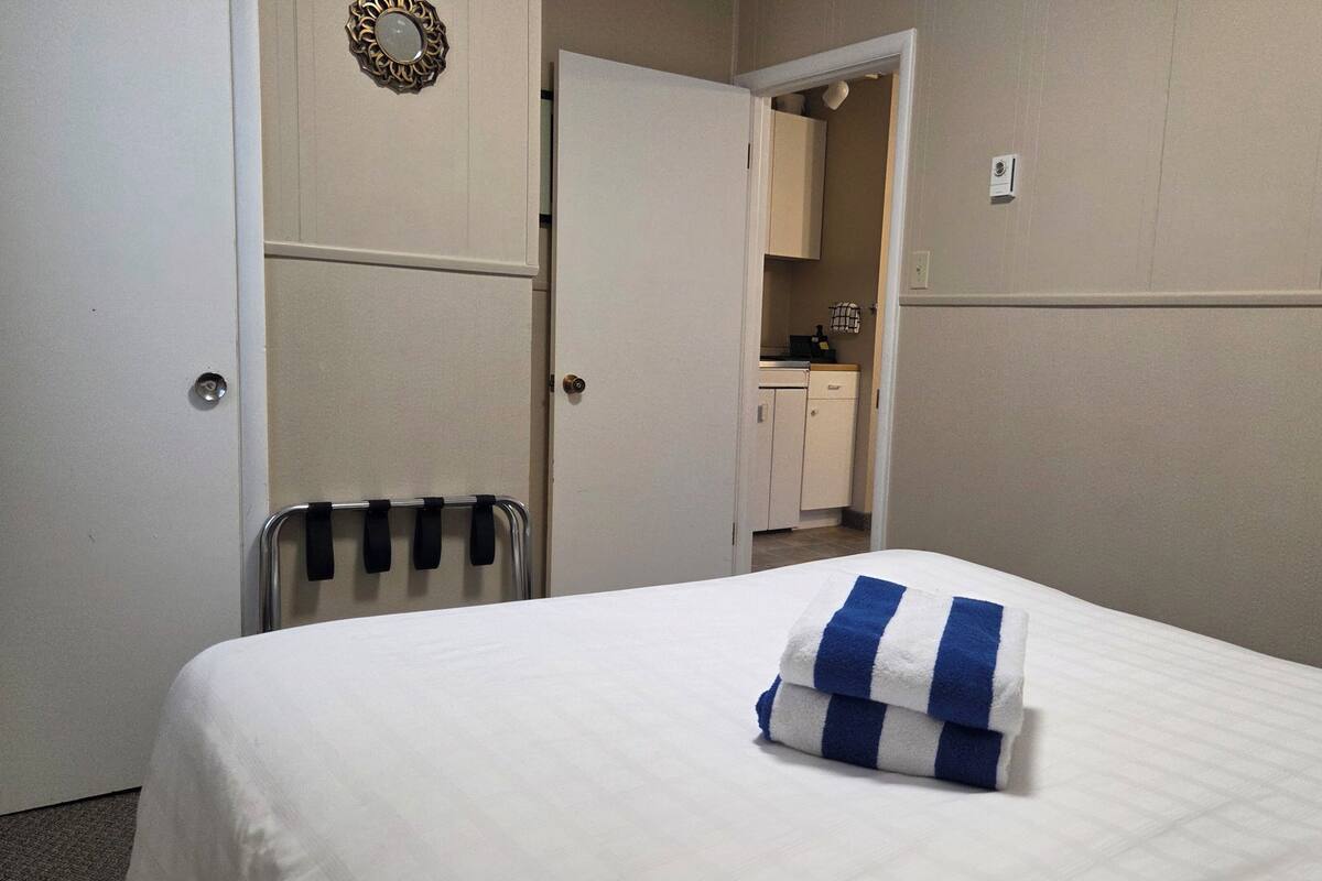 A hotel room with a bed, a chair, a door, and a kitchenette in the background.