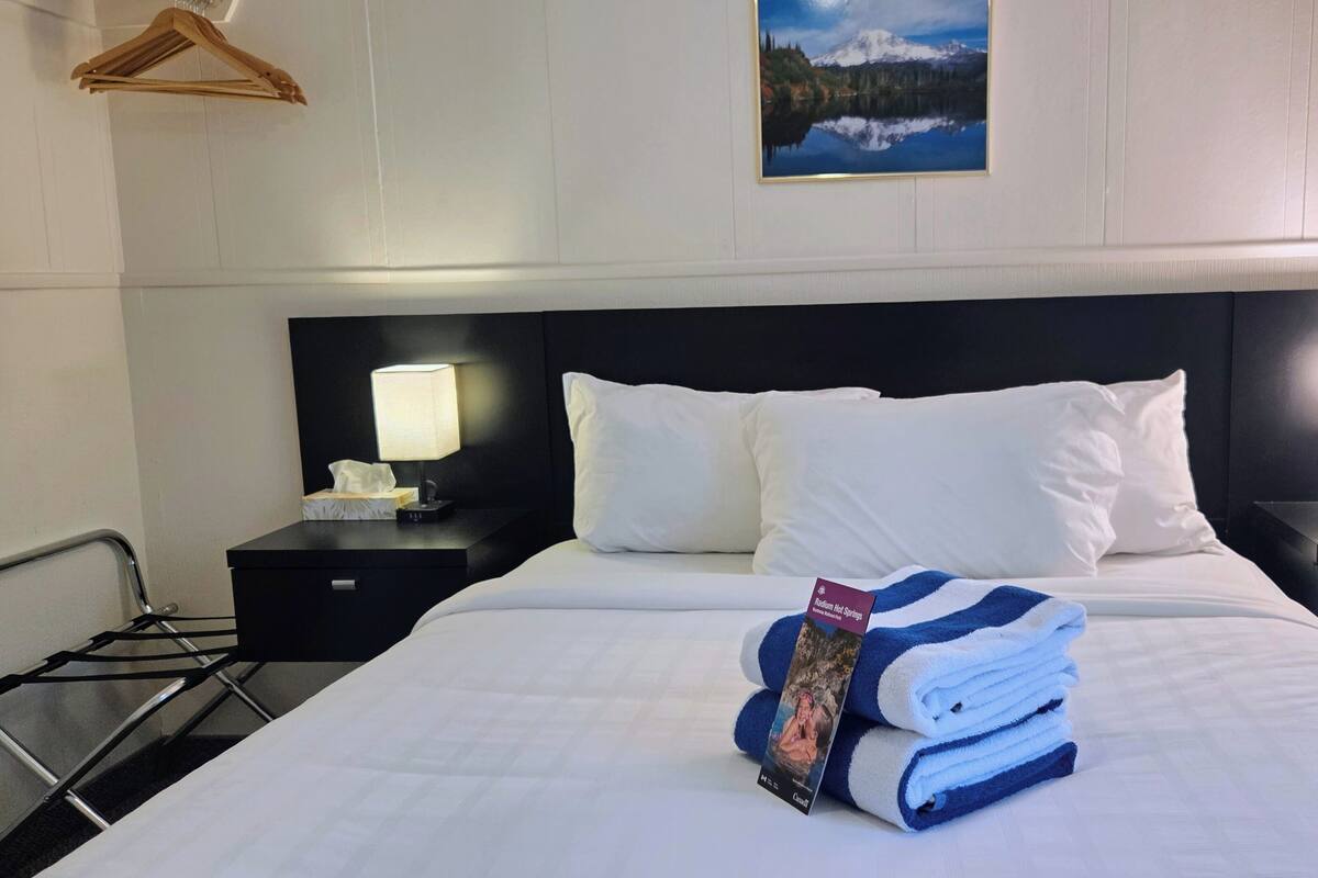 A hotel room with a bed, two bedside tables with lamps, a wooden hanger, a framed picture on the wall, and a folded towel with a magazine on the bed.