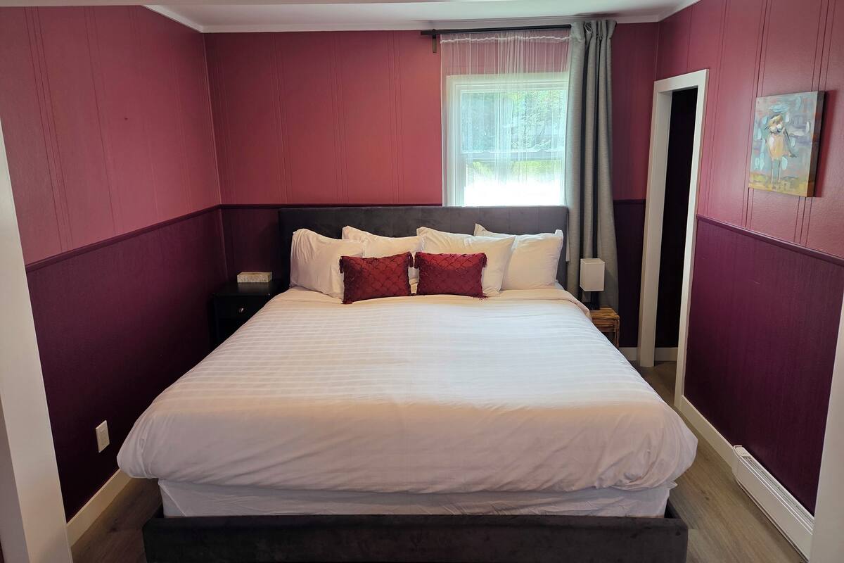 A neatly made bed with white and red pillows in a room with red walls and a window with sheer curtains.