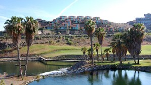 A golf course with a water feature, palm trees, and a large building with multiple balconies in the background. - Salobre Golf Villas - Holiday Rental Par 4 - 8 (San Bartolomé de Tirajana)