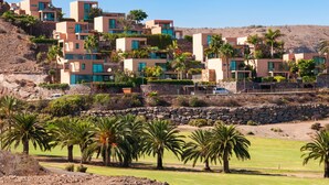 A hillside residential area with modern buildings, palm trees, and a stone wall. - Los Lagos 5- Holiday Rental Salobre Golf (San Bartolomé de Tirajana)