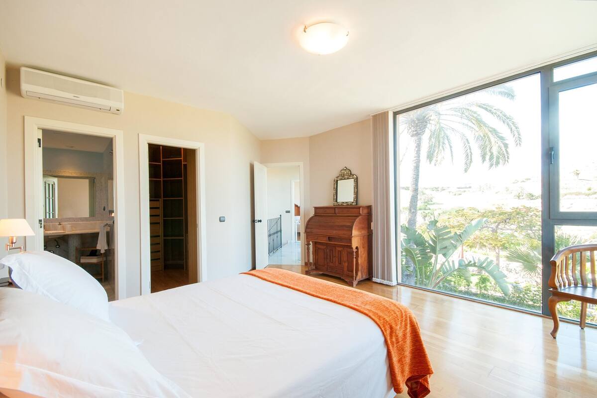 A hotel room with a bed, a wooden dresser, a mirror, and a large window with a view of palm trees.