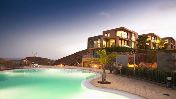 A swimming pool with a curved edge, surrounded by a paved walkway. There are palm trees and a lifebuoy near the pool. - Los Lagos 5- Holiday Rental Salobre Golf (San Bartolomé de Tirajana)