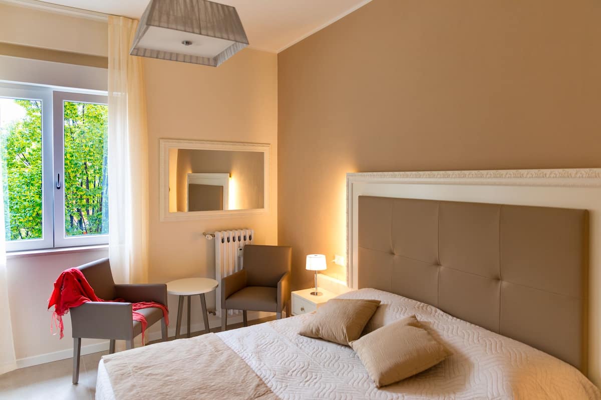 A hotel room with a large bed, a chair, a small table, a lamp, and a window with a view of trees.