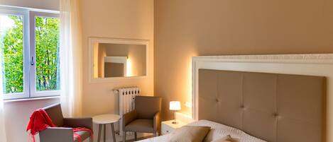 Hotelkamer met een groot bed, een stoel, een kleine tafel, een lamp en een raam met uitzicht op bomen.
