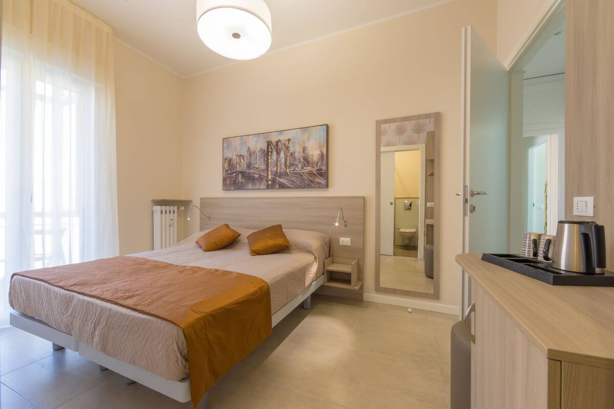 A modern hotel room with a large bed, bedside tables, a mirror, and a view of the cityscape through the window.