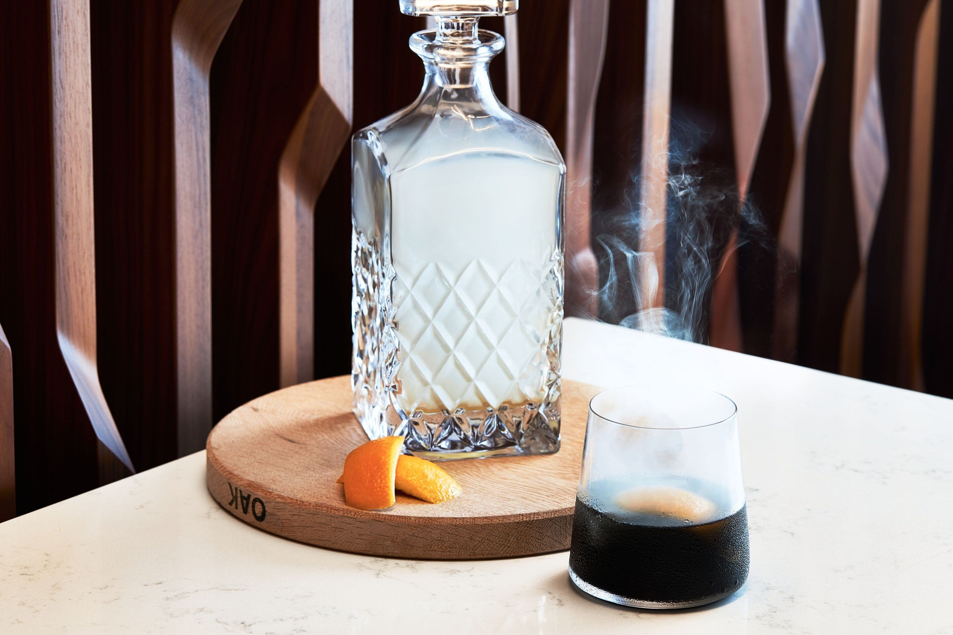 A clear glass decanter on a wooden coaster, a glass of dark liquid, and a half orange on the table.