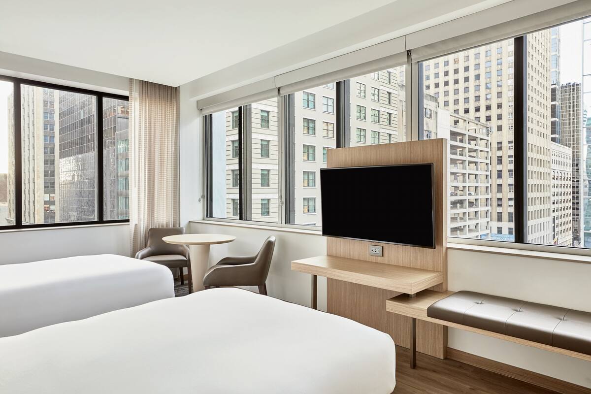 A modern hotel room with a large window, a flat-screen TV, a bed, a desk, and a chair.