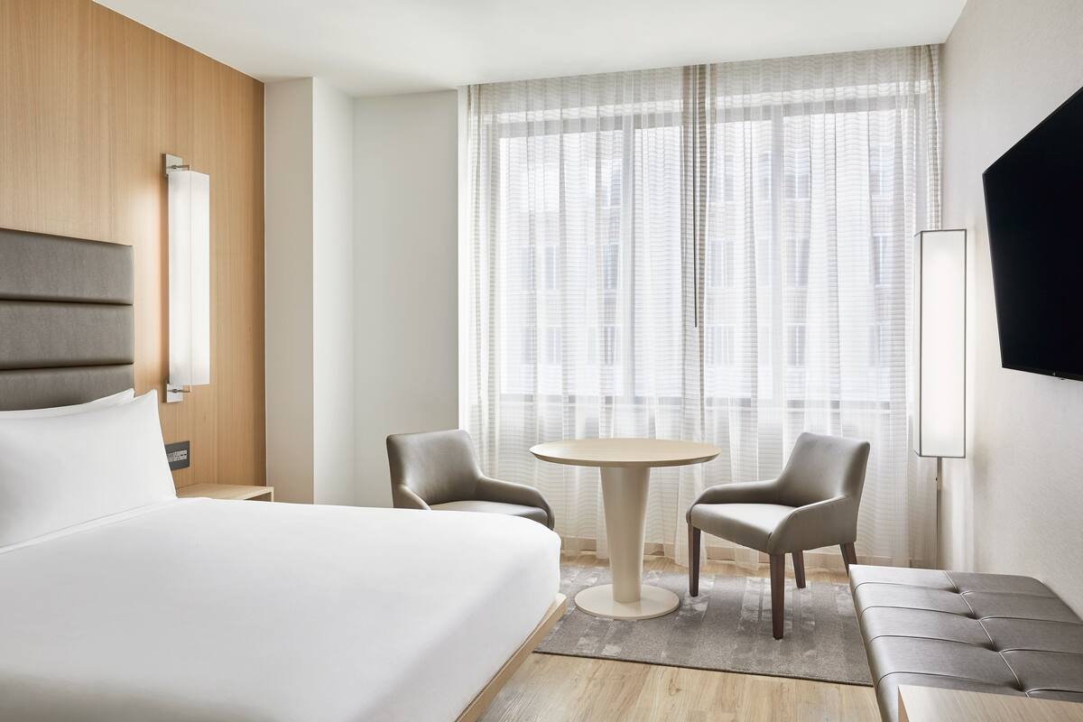 A hotel room with a large bed, a round table, two chairs, a TV, and a window with sheer curtains.