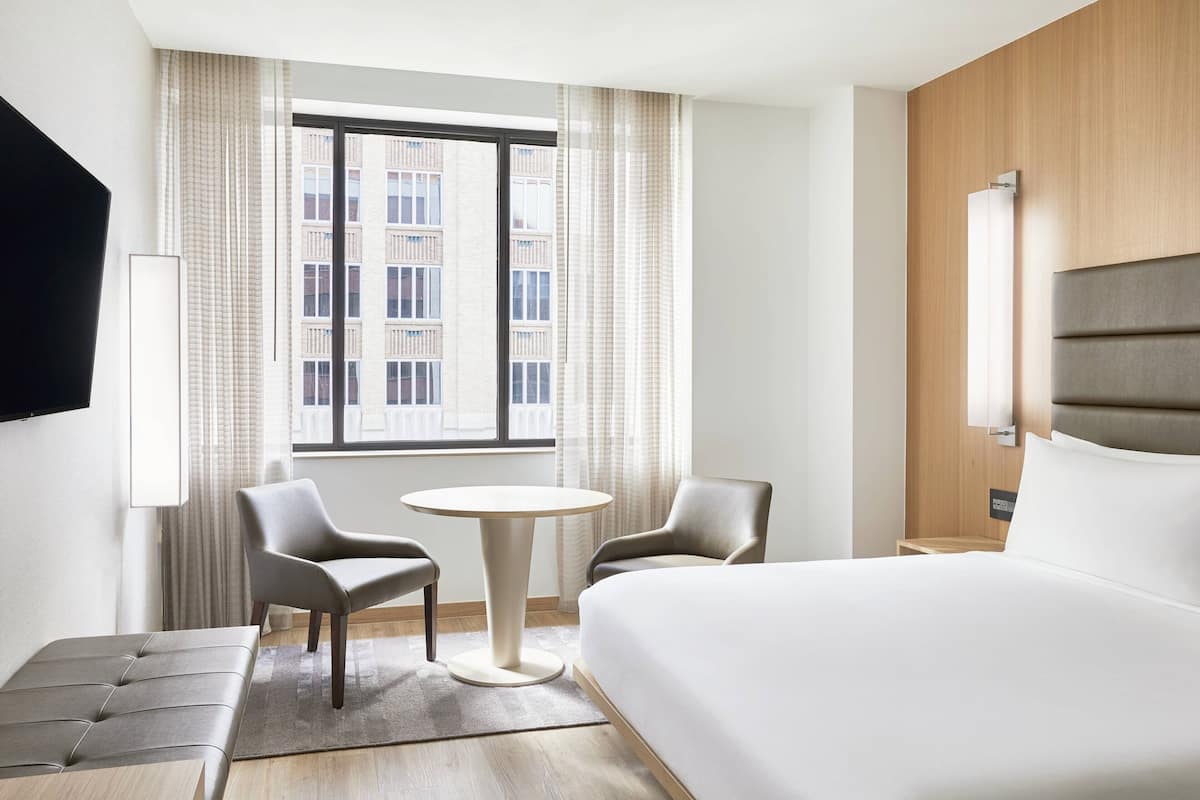 A hotel room with a large bed, two chairs, a small round table, and a TV mounted on the wall.