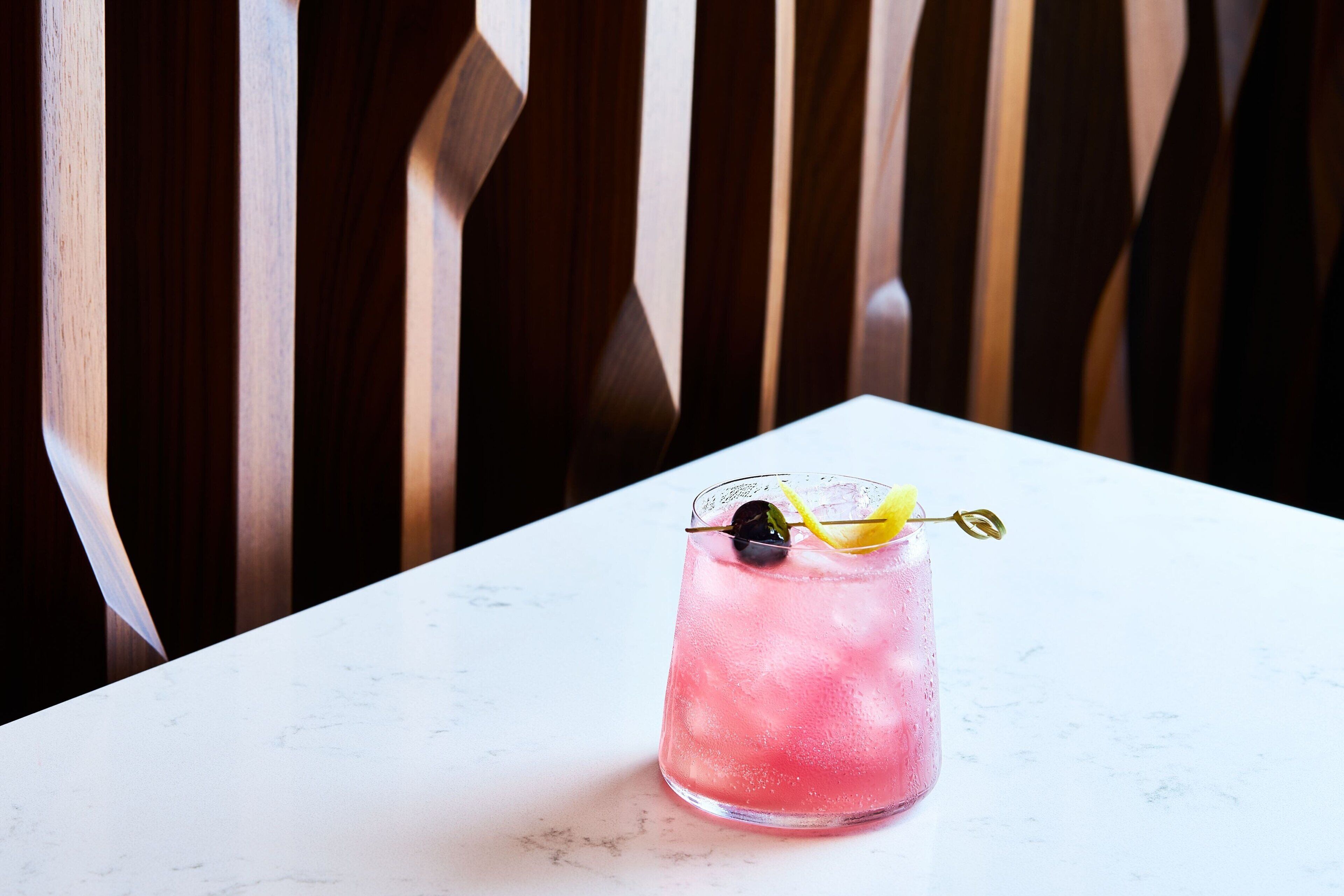 A pink beverage in a glass with a lemon slice and a gold straw on a white marble table.