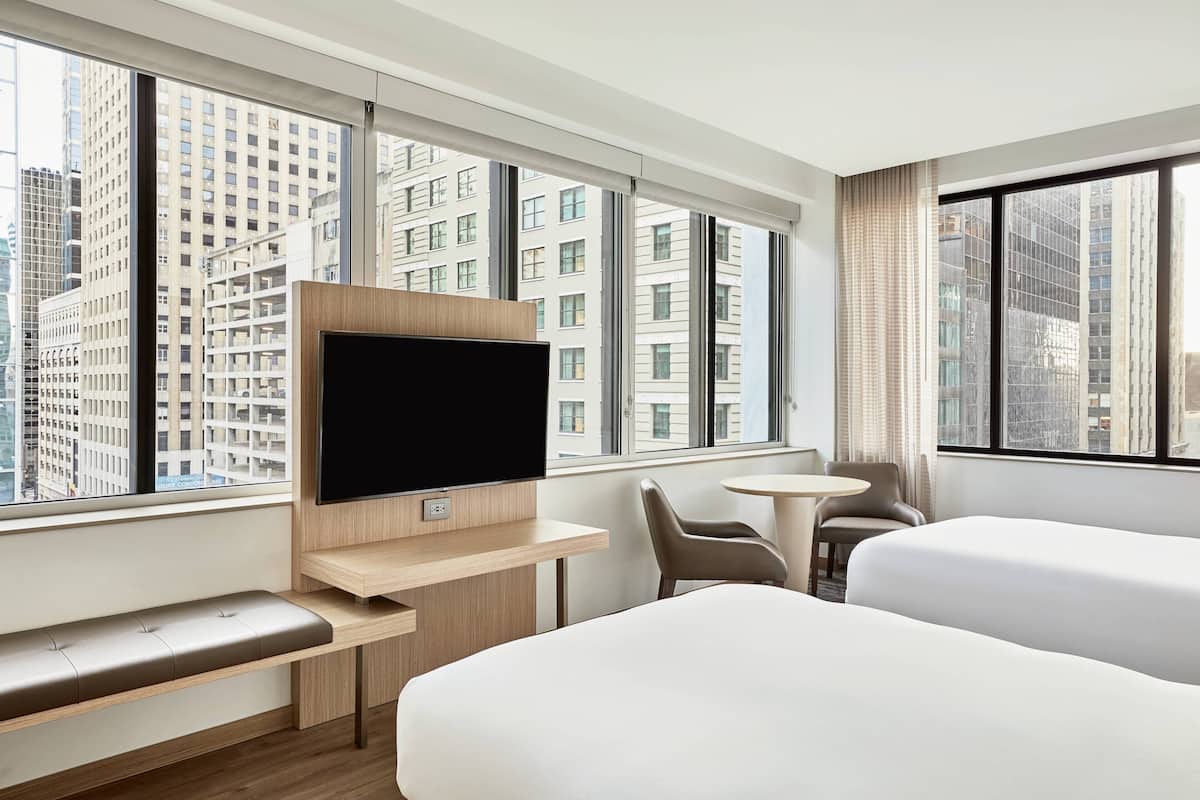 A modern hotel room with a large window, a flat-screen TV, a bench, a small round table, and two beds.