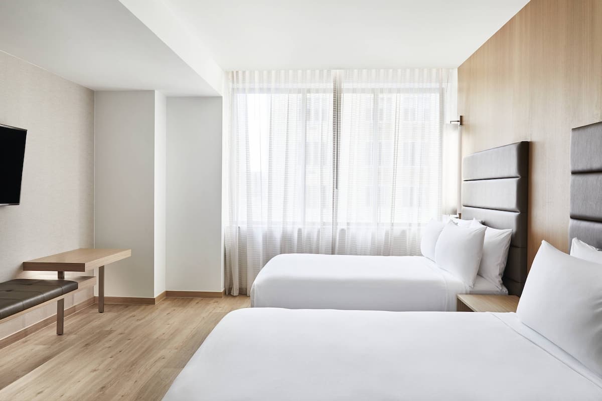 A modern hotel room with two beds, a wooden bench, a flat-screen TV, and a large window with sheer curtains.